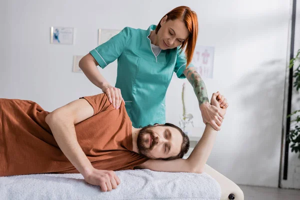 Redhead Rehabilitologist Massaging Painful Shoulder Bearded Man Closed Eyes Consulting — Stock Photo, Image