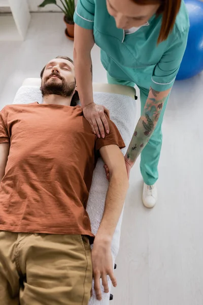 在康复中心 大胡子男子躺在理疗师身边进行胳膊和肩膀按摩的高视角图像 — 图库照片