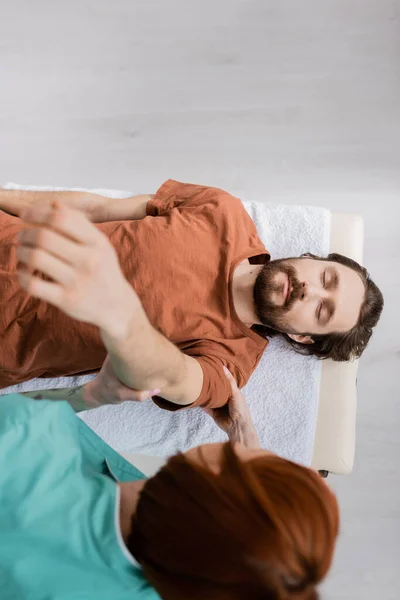 在会客室按摩床上闭锁的胡须男子伸开受伤的手臂 俯瞰模糊的脊椎按摩师 — 图库照片
