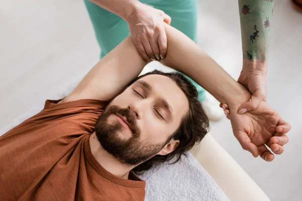 理学療法士の近くに目を閉じた髭の男が腕マッサージをしている様子を診療所でリハビリ中 — ストック写真