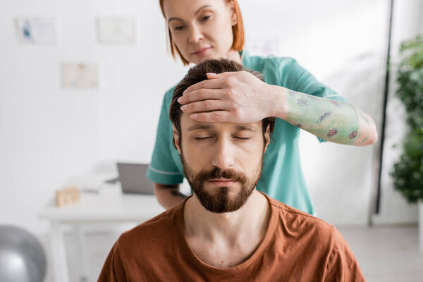 blurred physiotherapist touching forehead of bearded man with closed eyes during diagnostics in consulting room