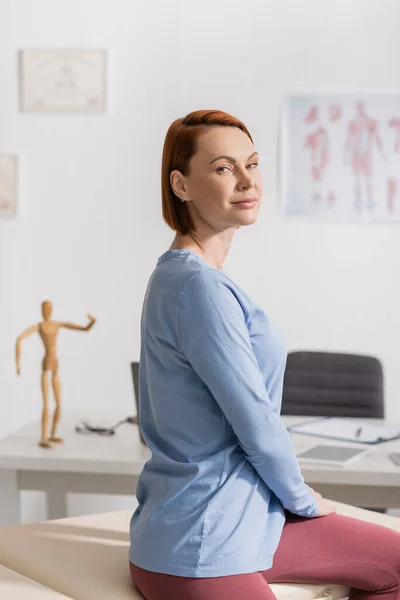 stock image pleased redhead woman smiling at camera while sitting in consulting room of rehabilitation center