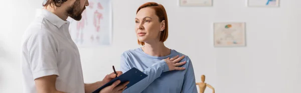 妇女摸着受伤的肩部 靠近理疗师 在诊疗室的剪贴板上写下诊断 — 图库照片