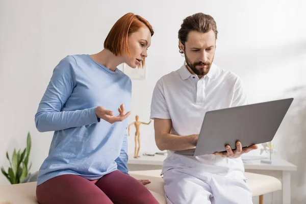Bearded Physiotherapist Using Laptop Redhead Woman Pointing Hand Consulting Room — Stock Photo, Image