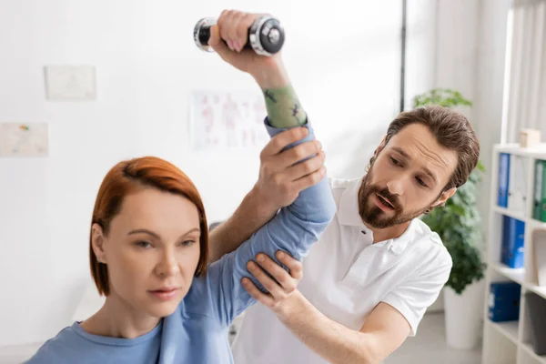 リハビリテーションセンターでダンベルを使って運動する赤毛の女性の腕を支える髭の理学療法士 — ストック写真
