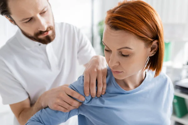 診察室で診断中に負傷した赤毛の女性の肩に触れる理学療法士 — ストック写真