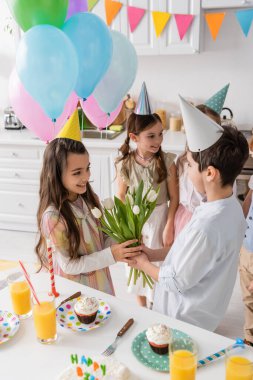 preteen boy giving tulips to cheerful birthday girl near kids on blurred background  clipart