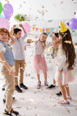 Parti şapkalı mutlu çocuklar evde doğum günü kutlaması sırasında konfeti altında dans ediyorlar. 