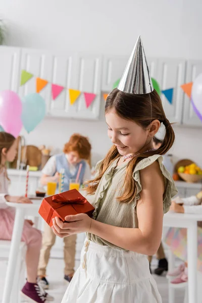 Szczęśliwa Dziewczyna Party Cap Gospodarstwa Prezent Urodzinowy Blisko Przyjaciół Zamazanym — Zdjęcie stockowe