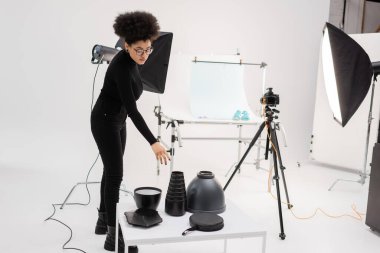 Afro-Amerikan içerik üreticisi fotoğraf stüdyosundaki tripodda ışık ekipmanlarına, yansıtıcı ve dijital kameraya uzanıyor.