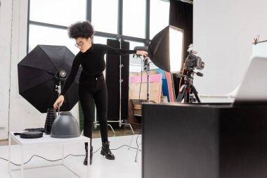 african american content producer in black clothes reaching lamps near softbox reflector and digital camera in modern photo studio clipart