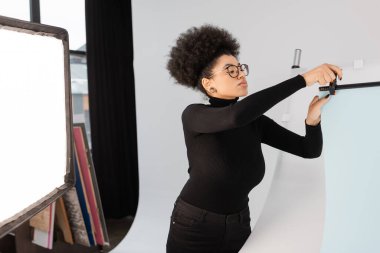 Gözlüklü Afro-Amerikan içerik yapımcısı fotoğraf stüdyosunda yansıtıcının yanına arka plan sayfası kuruyor.