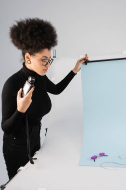 Afro-Amerikan içerik üreticisi beyaz arka plan örtüsünün yanında duruyor ve çekim masasında son moda güneş gözlükleri var.