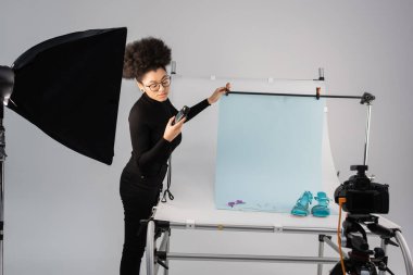 Afro-Amerikalı içerik yapımcısı yansıtıcı ve fotoğraf stüdyosunda moda ayakkabılar ve güneş gözlükleriyle çekim masasına bakıyor.