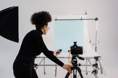 african american content producer holding exposure meter near digital camera and shooting table in modern studio clipart