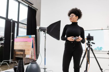 Afro-Amerikan içerik prodüktörü fotoğraf stüdyosundaki softbox yansıtıcı ve dijital kameranın yanından uzağa bakıyor.