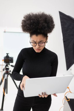 Rahatsız ve somurtkan Afro-Amerikan içerik üreticisi fotoğraf stüdyosundaki bulanık dijital kameranın yanında laptop kullanıyor.