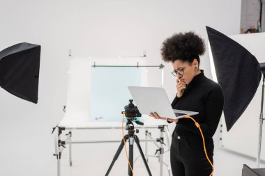 Düşünceli Afro-Amerikan içerik yöneticisi modern fotoğraf stüdyosundaki spot ışıkları ve dijital kameranın yanındaki dizüstü bilgisayara bakıyor.