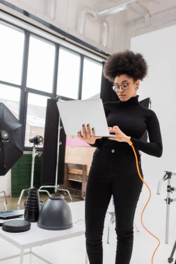 african american content manager in eyeglasses using laptop while standing near lighting equipment in photo studio clipart