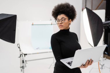 Afro-Amerikan içerik yöneticisi dizüstü bilgisayarla spot ışıklarının yakınına bakıyor ve fotoğraf stüdyosundaki bulanık çekim masasına bakıyor.