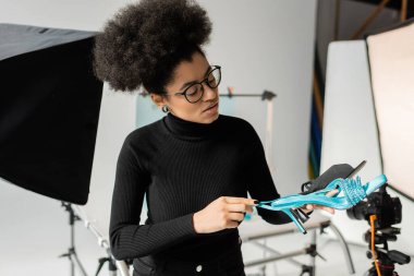Siyah balıkçı yaka ve gözlüklü Afro-Amerikan içerik üreticisi bulanık fotoğraf stüdyosundaki şık ayakkabılara bakıyor.