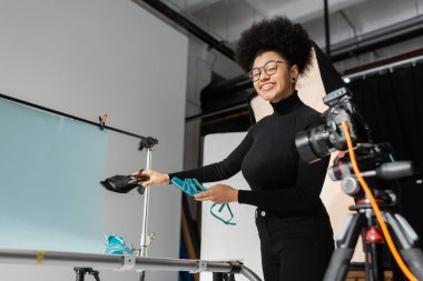 Neşeli Afro-Amerikan içerik yapımcısı fotoğraf stüdyosundaki son moda ayakkabıları elinde tutarken kameraya bakıyor.