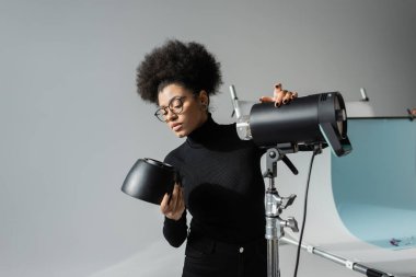 Siyah balıkçı yaka kazaklı ve gözlüklü Afro-Amerikan içerik üreticisi fotoğraf stüdyosunda flaş lambası detaylarını saklıyor.