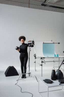 full length of happy african american content maker near strobe lamp and shooting table in contemporary photo studio clipart