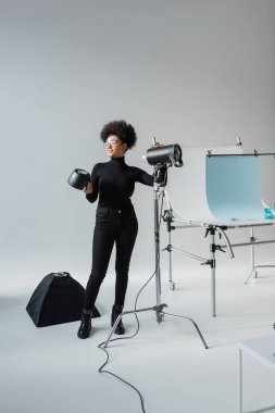 Çekim masasının yanında fotoğraf stüdyosunda ışıklandırma ekipmanı toplayan Afro-Amerikan içerikli tam boy bir yapımcı.