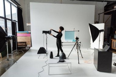 full length of african american content producer in black clothes assembling lighting equipment in spacious photo studio clipart