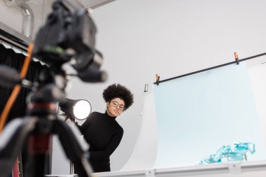 Afro-Amerikan içerik üreticisi spot ışıklarının yanındaki bulanık dijital kameraya bakıyor ve fotoğraf stüdyosunda moda ayakkabıları olan bir masa çekiyor.