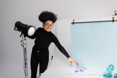 Mutlu Afro-Amerikan içerik yapımcısı flaş lambanın yanındaki kameraya bakıyor ve fotoğraf stüdyosunda son moda güneş gözlükleri ve ayakkabılarla çekim masasına oturuyor.