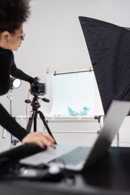 african american content maker adjusting digital camera near shooting table with trendy shoes and blurred laptop in photo studio clipart