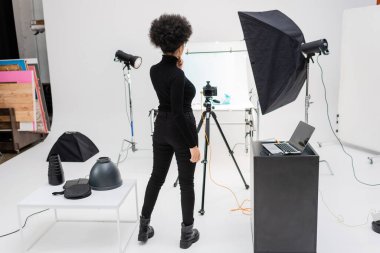 Siyah elbiseli Afro-Amerikan içerik yöneticisinin arka görüntüsü dizüstü bilgisayarın yanında duruyor ve fotoğraf stüdyosunda tripodun üzerinde dijital kamera var.