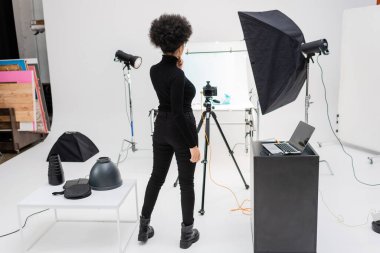 back view of african american content manager in black clothes standing near laptop and digital camera on tripod in photo studio clipart