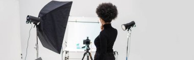 Afro-Amerikan içerik yöneticisinin arka görüntüsü siyah kıyafetler içinde ışıklandırma ekipmanı ve dijital kamera ile fotoğraf stüdyosunda, afiş
