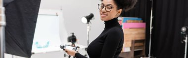 Pozitif Afro-Amerikalı gözlüklü içerik üreticisi elinde fotoğraf stüdyosundaki fotoğraf makinesine gülümsüyor.