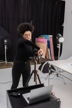 Gözlüklü Afro-Amerikan içerik yapımcısı fotoğraf stüdyosunda dizüstü bilgisayarın yanında dijital kamera monte ediyor.
