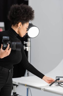 Afro-Amerikan içerik yapımcısı dijital kamerasıyla kozmetik fırçasını fotoğraf stüdyosundaki çekim masasında tutuyor.