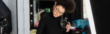 african american photographer in eyeglasses holding professional digital camera in photo studio, banner clipart