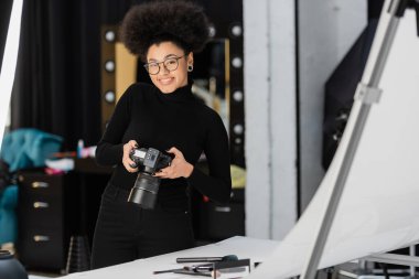 Fotoğraf stüdyosunda dekoratif kozmetik ürünlerinin yanında dijital kamerayla gülümseyen neşeli Afro-Amerikan içerik yapımcısı.