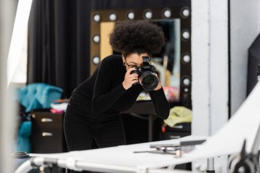 african american content maker taking photo of decorative cosmetics on blurred shooting table in photo studio clipart