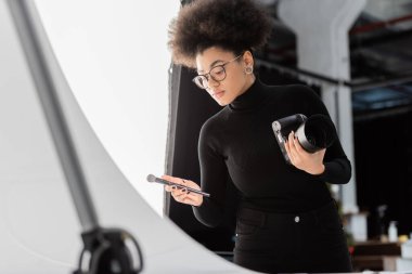 Afro-Amerikan içerik yöneticisi fotoğraf stüdyosunda dijital kamera ve kozmetik fırça tutuyor.
