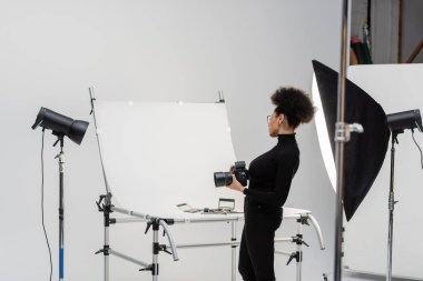 Fotoğraf stüdyosundaki dekoratif kozmetik ve güzellik aletlerine bakan dijital kameralı Afro-Amerikan içerik üreticisinin yan görüntüsü