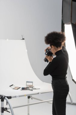 Siyah balıkçı yakalı Afrikalı Amerikalı fotoğrafçı fotoğraf stüdyosundaki dekoratif kozmetik ürünlerinin fotoğrafını çekiyor.