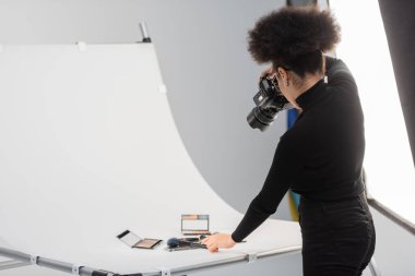 Afro-Amerikan fotoğrafçı, fotoğraf stüdyosundaki çekim masasında güzellik aletleri ve dekoratif kozmetik ürünlerinin fotoğrafını çekiyor.