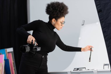 stylish african american content producer holding cosmetic brush near decorative cosmetics on shooting table in photo studio clipart