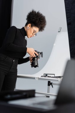 african american photographer in black turtleneck and eyeglasses looking at digital camera near decorative cosmetics in photo studio clipart