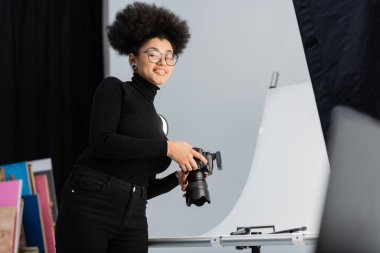 Gözlüklü neşeli Afrikalı Amerikalı fotoğrafçı fotoğraf stüdyosundaki dekoratif kozmetik ve güzellik aletlerinin yanındaki kameraya gülümsüyor.