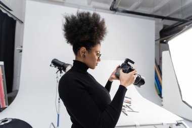 Siyah balıkçı yaka kazaklı Afro-Amerikan içerik üreticisinin fotoğraf stüdyosundaki dijital kameraya bakışının yan görüntüsü.